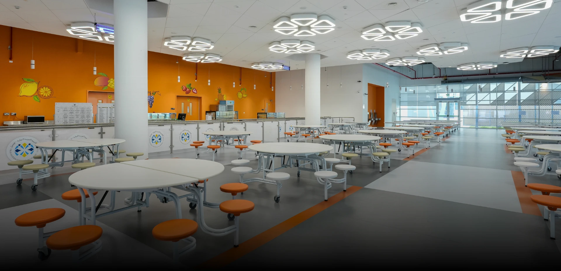 A brightly designed school cafeteria by an interior design and fitout company, featuring orange walls with fruit decals, white round tables with attached orange stools, and unique ceiling lights for learning spaces.