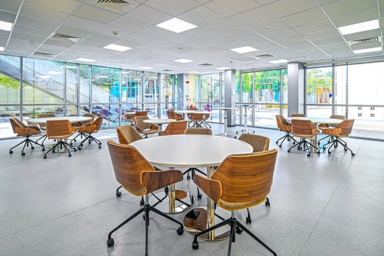 A modern corporate office meeting area with circular white tables, wooden and grey chairs, and large windows overlooking a city, designed as functional workspaces.