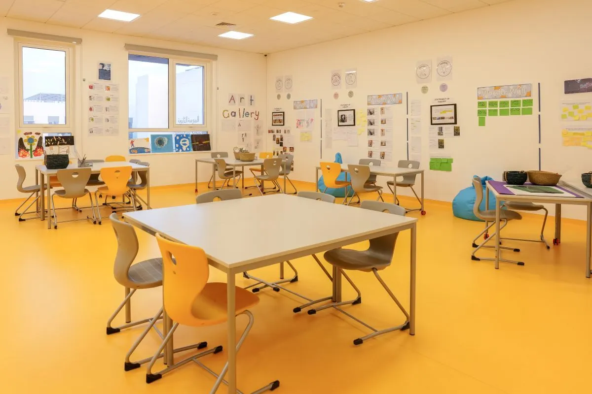 A bright architectural learning space with a yellow floor, numerous tables and chairs, and walls adorned with student artwork and displays, characteristic of an art classroom.