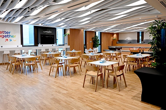 A spacious corporate office cafe and breakout area with wavy ceiling panels, white and wooden furniture, a tiered seating area, and a foosball table, epitomizing modern workspaces.