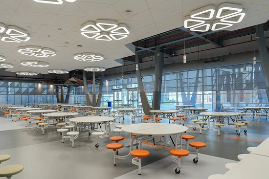 A large, modern school cafeteria designed by an interior design and fitout company, with round white tables, attached orange stools, and expansive windows, ideal for learning spaces.