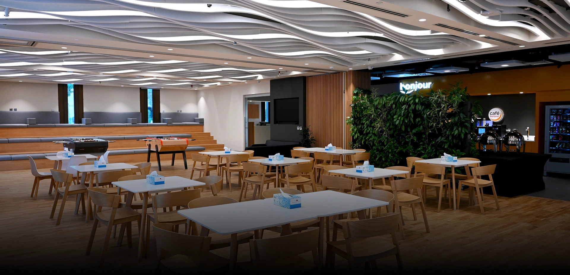 A vibrant office café and breakout area, designed by an interior design and fit out company, with white tables, wooden chairs, a tiered seating area, and a green wall.
